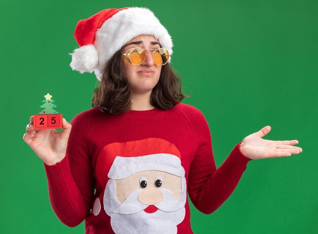 サンタの帽子をかぶったクリスマスセーターの少女と、緑の壁の上に立って腕を上げて混乱している25番のおもちゃの立方体を持っている眼鏡
