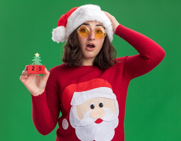 Young girl in christmas sweater wearing santa hat and glasses holding toy cubes with number twenty five  surprised and amazed standing over green wall