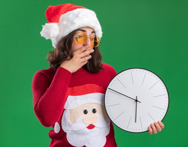 Young girl in christmas sweater wearing santa hat and glasses holding wall clock looking at it amazed and surprised covering mouth with hand standing over green background