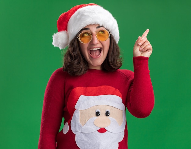 サンタの帽子と眼鏡をかけて幸せで陽気な人差し指を緑の壁の上に立って見上げるクリスマスセーターの少女