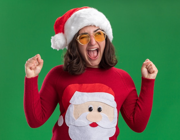 Free photo young girl in christmas sweater wearing santa hat and glasses shouting excited and crazy happy clenching fists standing over green wall