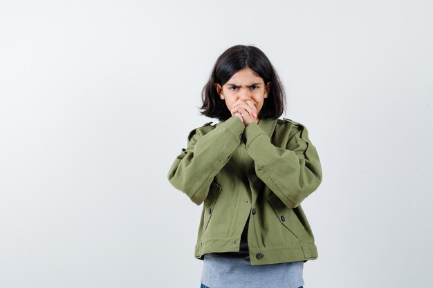 Young girl clasping hands in grey sweater, khaki jacket, jean pant and looking angry. front view.