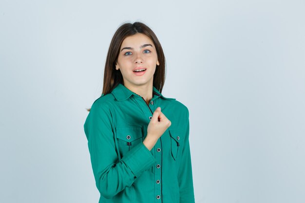 Young girl clenching fist over chest in green blouse and looking cheery. front view.