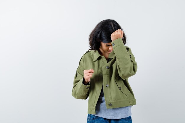 Young girl clenching fist and putting hand on head in grey sweater, khaki jacket, jean pant and looking tired. front view.