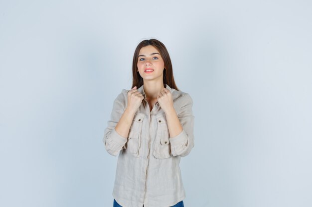 Young girl clenching fists over chest in beige shirt, jeans and looking cheery. front view.