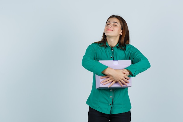 Young girl clutching gift box to her chest in green blouse, black pants and looking merry. front view.