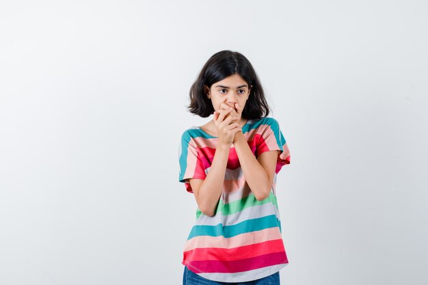Young girl in colorful striped t-shirt covering mouth with hands and looking surprised , front view.