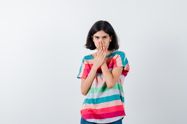 Young girl in colorful striped t-shirt covering mouth with hands and looking timid , front view.