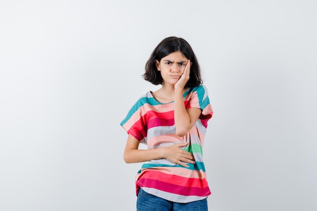 Young girl in colorful striped t-shirt having toothache and looking annoyed , front view.