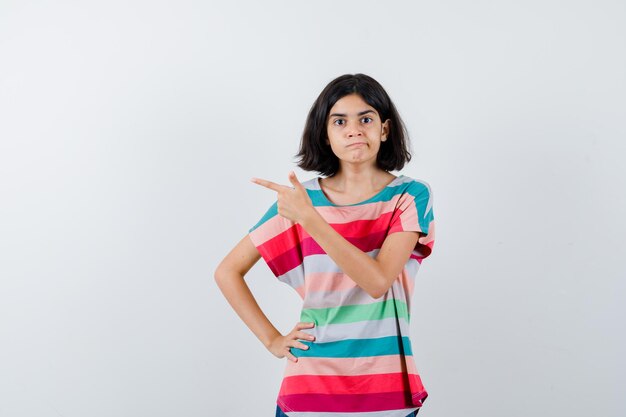 Young girl in colorful striped t-shirt pointing left with index finger while holding hand on waist and looking serious , front view.