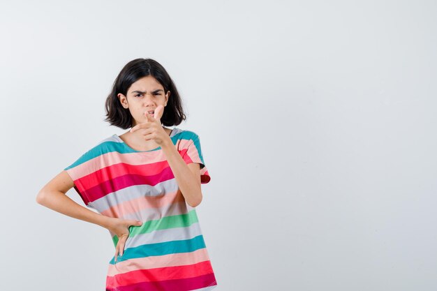 Young girl in colorful striped t-shirt pointing  while holding hand on waist and looking angry , front view.