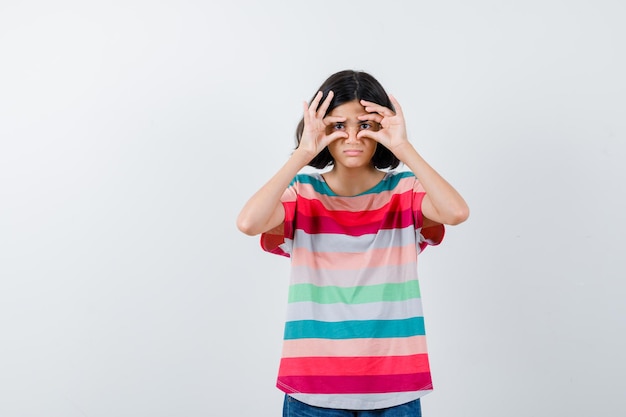 Young girl in colorful striped t-shirt showing binoculars gesture and looking cute , front view.