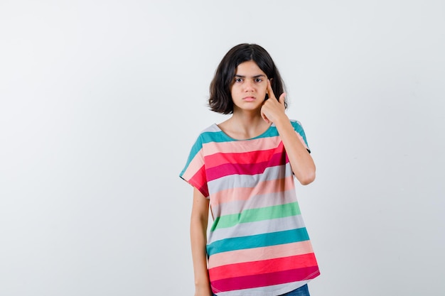 Giovane ragazza in maglietta a righe colorata che mostra il gesto della pistola vicino alla testa e sembra seria, vista frontale.