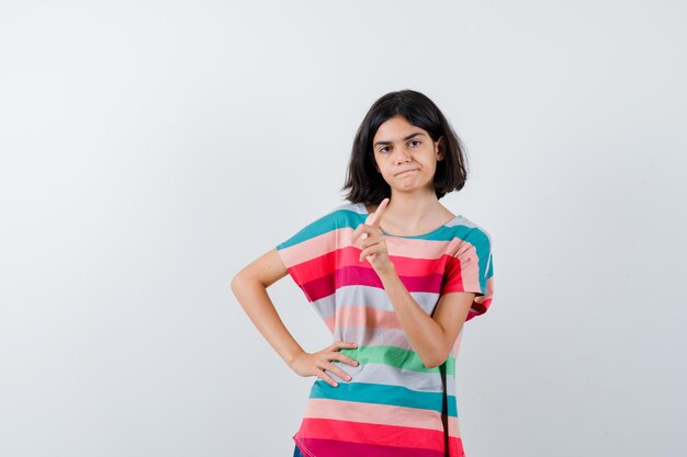 Young girl in colorful striped t-shirt showing warning gesture while holding hand on waist and looking serious , front view.