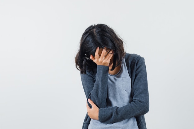 Free photo young girl covering face with hand in light gray t-shirt and dark grey zip-front hoodie and looking stressed.