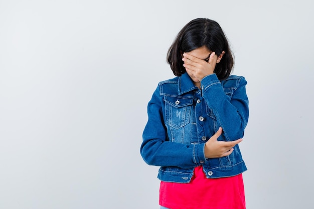 Young girl covering face with hand in red t-shirt and jean jacket and looking harried. front view.