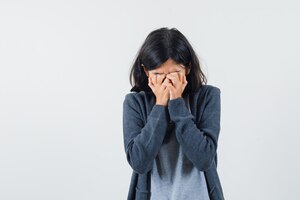 Young girl covering face with hands in light gray t-shirt and dark grey zip-front hoodie and looking sad.