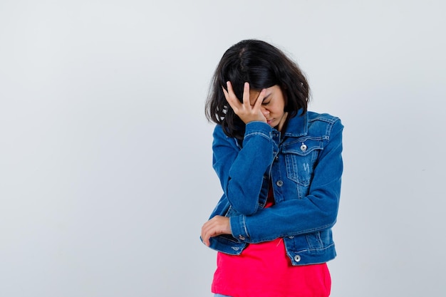 Young girl covering forehead with hand in red t-shirt and jean jacket and looking tired. 