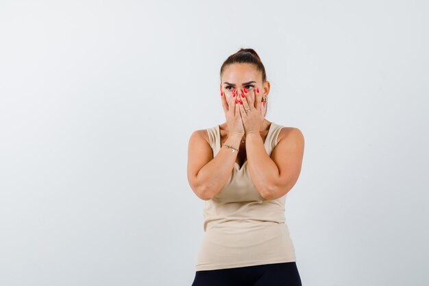 Young girl covering mouth and nose with hands in beige top, black pants and looking excited , front view.