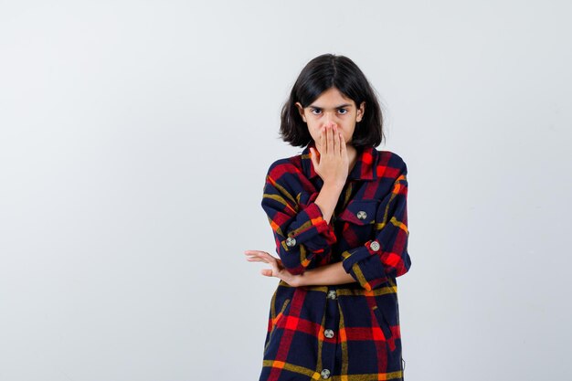 Young girl covering mouth with hand in checked shirt and looking serious. front view.
