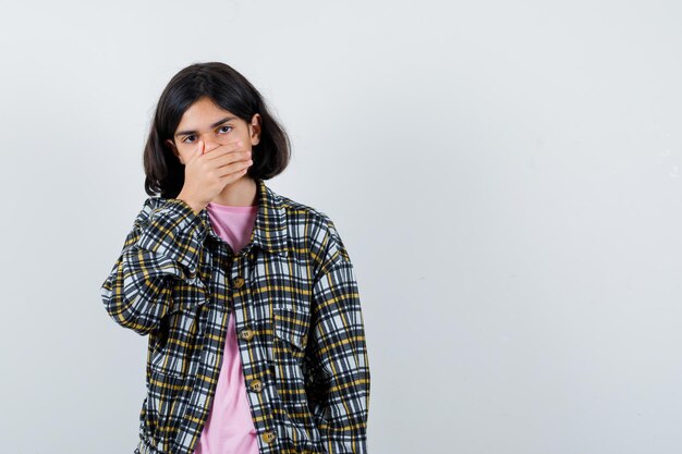 チェックシャツとピンクのTシャツで手で口を覆い、臆病に見える少女。正面図。