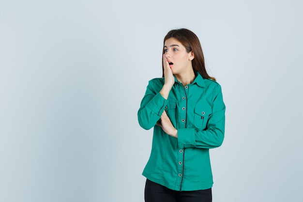 Young girl covering mouth with hand, keeping mouth wide open in green blouse, black pants and looking shocked. front view.