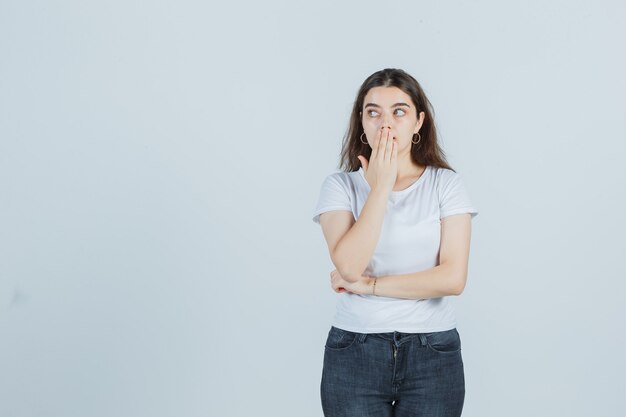 Young girl covering mouth with hand, looking aside in t-shirt, jeans and looking surprised. front view.