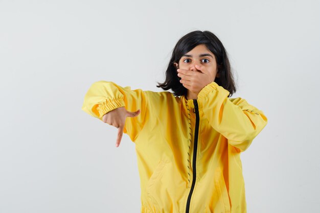 Молодая девушка закрывает рот рукой, указывает вниз указательным пальцем в желтой куртке-бомбардировщике и выглядит удивленно.