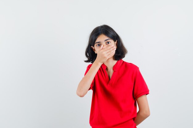Young girl covering mouth with hand in red t-shirt and looking surprised. front view.
