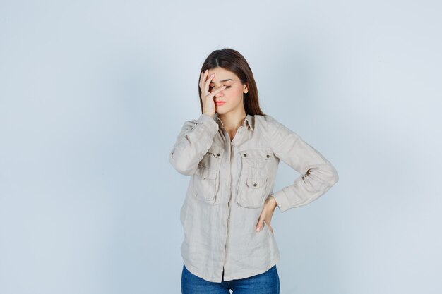 Young girl covering part of face with hand, with hand on hip in beige shirt, jeans and looking harried , front view.