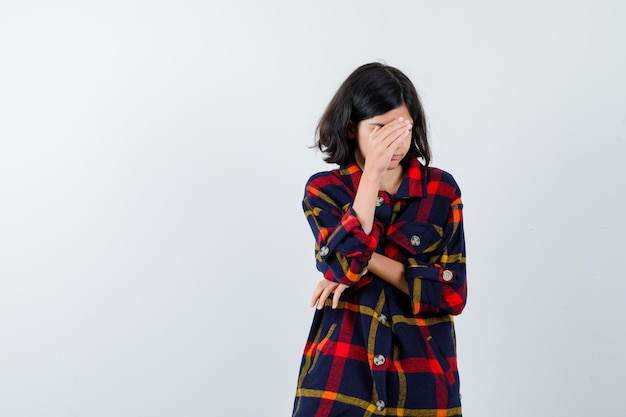 Young girl covering part of face with hands in checked shirt and looking tired , front view.