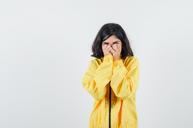 노란색 폭격기 재킷에 손으로 얼굴의 일부를 덮고 소심한 어린 소녀.