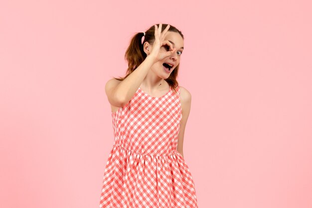 Free photo young girl in cute pink dress looking through her fingers on pink