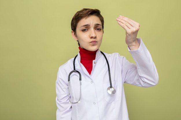 Молодая девушка-врач в белом халате со стетоскопом на шее смущена, делая деньги жестом, потирая пальцы, стоя на зеленом Бесплатные Фотографии