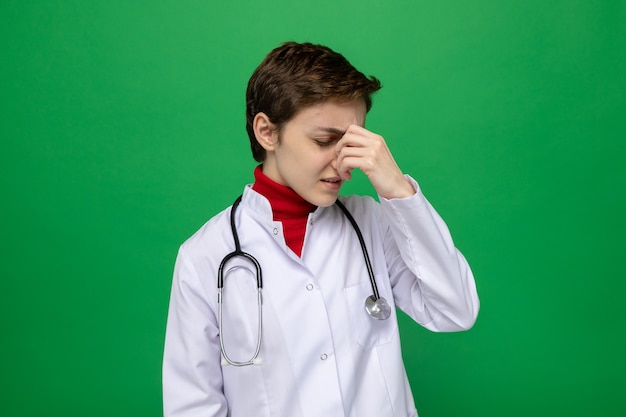 Молодая девушка-врач в белом халате со стетоскопом на шее выглядит нездоровой, усталой и напряженной, трогательно касаясь носа между закрытыми глазами, стоя на зеленом