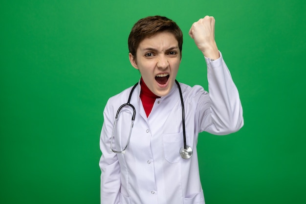 Молодая девушка-врач в белом халате со стетоскопом на шее кричит с агрессивным выражением лица, поднимая кулак, стоя над зеленой стеной Premium Фотографии