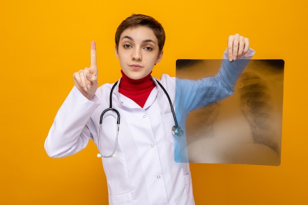 흰색 코트를 입은 어린 소녀 의사가 주황색 벽 위에 서 있는 검지 손가락을 보여주는 진지한 자신감 있는 표정으로 정면을 바라보는 폐 엑스레이를 들고 청진기를 들고 있습니다.