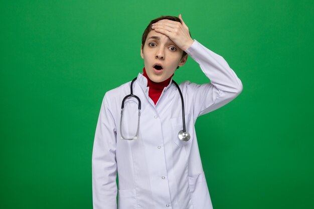 Young girl doctor in white coat with stethoscope around neck confused and worried holding hand on her forehead standing on green