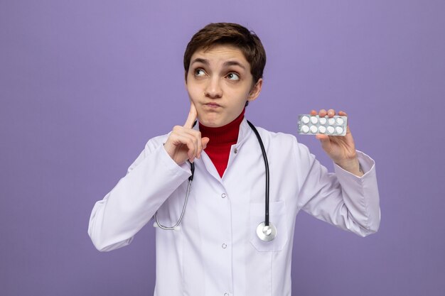 首の周りに聴診器を持った白いコートを着た若い女の子の医者は、紫色の壁の上に立って困惑して見上げる丸薬で水ぶくれを保持しています