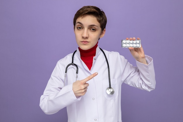 Medico della ragazza in camice bianco con lo stetoscopio intorno al collo che tiene la bolla con le pillole che indicano con il dito indice con la faccia seria che sta sopra il muro viola