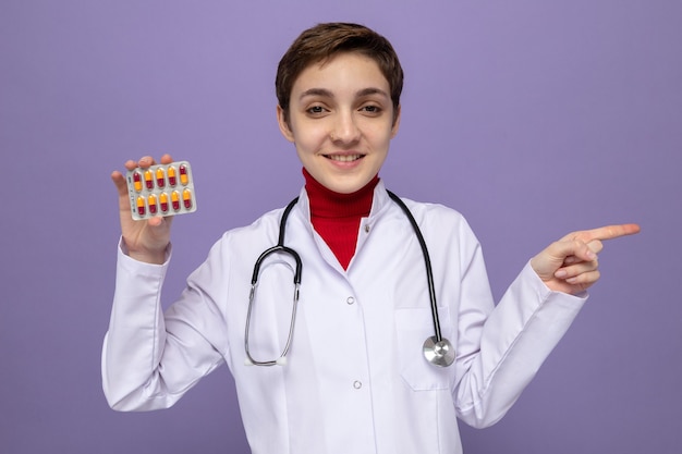 Free photo young girl doctor in white coat with stethoscope around neck holding blister with pills smiling cheerfully pointing with index finger to the side standing on purple