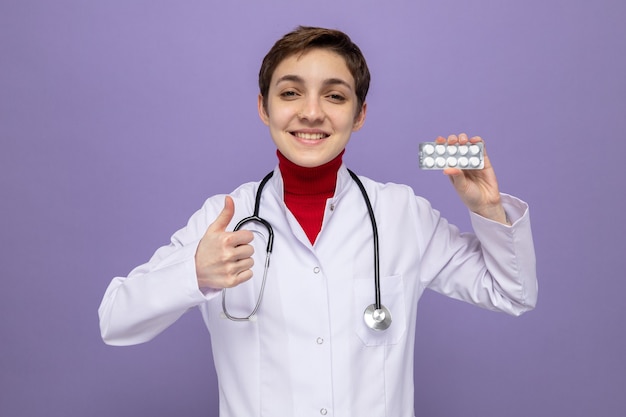 Free photo young girl doctor in white coat with stethoscope around neck holding blister with pills smiling cheerfully showing thumbs up standing on purple