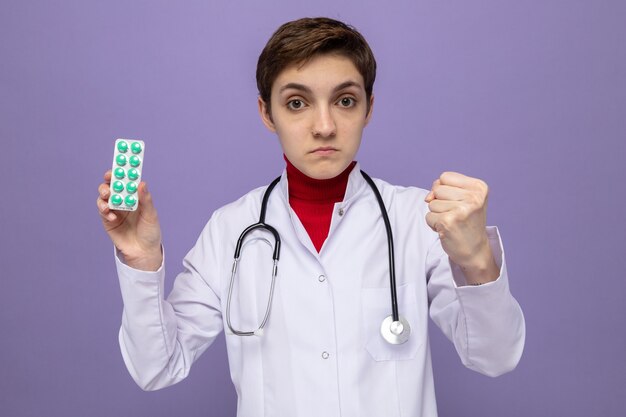 Young girl doctor in white coat with stethoscope around neck holding blister with pills  with serious face showing fist standing over purple wall