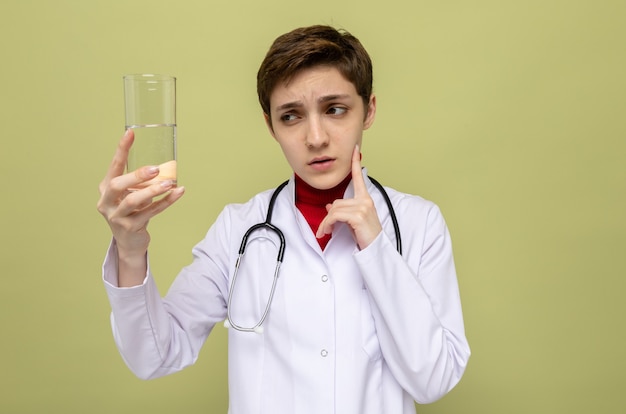 Молодая девушка-врач в белом халате со стетоскопом на шее держит стакан воды, озадаченный, стоя на зеленом