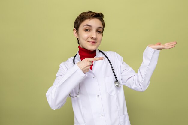 首の周りに聴診器を持った白いコートを着た若い女の子の医者は、彼女の手の腕を提示する側に人差し指で指している自信を持って笑顔に見えます