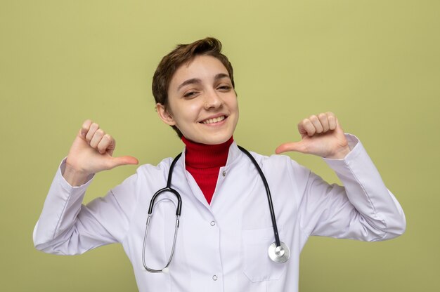 Young girl doctor in white coat with stethoscope around neck smiling confident pointing at herself standing on green