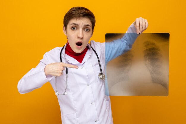 Young girl doctor in white coat with stethoscope holding x-ray of lungs pointing with index finger at it looking amazed and surprised standing on orange