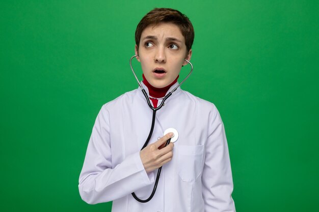 Young girl doctor in white coat with stethoscope listening to her heartbeat looking worried standing on green