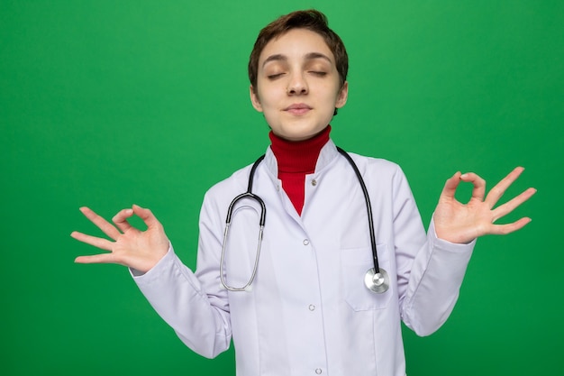 Foto gratuita medico della ragazza in camice bianco con lo stetoscopio che fa il gesto di meditazione che prova a rilassarsi con gli occhi chiusi