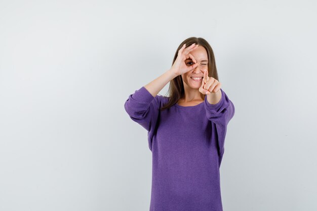 Young girl doing ok sign on eye and pointing at camera in violet shirt front view.